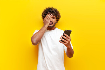 young indian sad guy using smartphone online with facepalm on yellow isolated background - obrazy, fototapety, plakaty