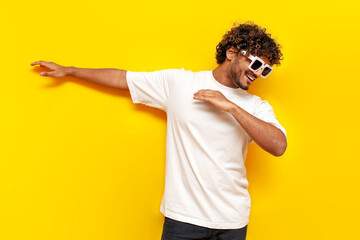 young indian guy in sunglasses dancing dab on yellow isolated background, south asian man in white...