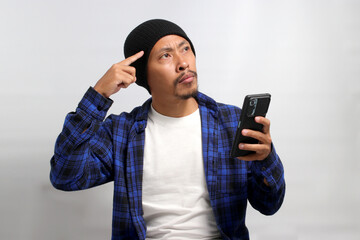 Pensive Asian man, dressed in a beanie hat and casual shirt, holds a mobile phone, appearing...