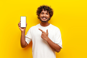 young indian guy pointing at blank smartphone screen on yellow isolated background, south asian man...