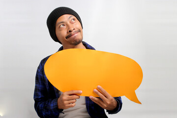 Pensive young Asian man in a beanie hat and casual shirt seems excited as he holds a speech bubble,...