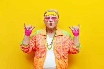 funny crazy old granny in sports hipster clothes shows youth gesture on yellow isolated background