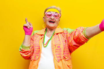 funny crazy old granny in sports hipster clothes takes selfie online and points on yellow isolated...