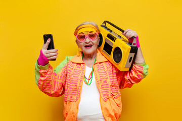 crazy old grandmother with tape recorder and smartphone in hipster clothes listens music on yellow background
