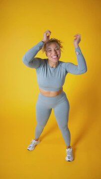 Vertical video studio shot looking down on full length shot of smiling woman wearing gym fitness clothing exercising and dancing on yellow background - shot in slow motion