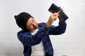 Shocked young Asian man, dressed in a beanie hat and casual shirt, looks angrily at his empty wallet, facing financial difficulties that might lead to bankruptcy, isolated on a white background