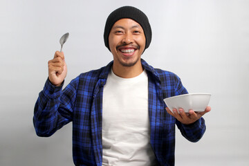 Smiling Asian man, clad in a beanie hat and casual shirt, holds a spoon and a bowl of cereal for...
