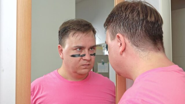 Man looks at reflection in mirror and applies black war paint in preparation for attack. Angry man applies war paint to struggle against criminals