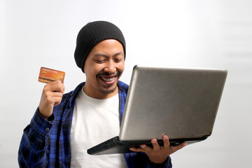 An excited young Asian man, dressed in a beanie hat and casual shirt, enthusiastically uses a...
