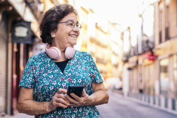 Old woman using smartphone and headphones walking down the street