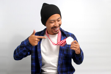 A proud young Asian man, dressed in a beanie hat and casual shirt, smiles as he points at his medal while standing against a white background