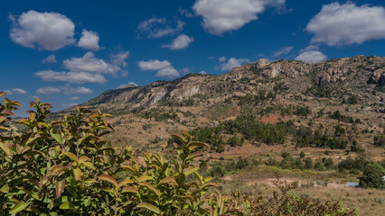 A mountain with rocky slopes against a background of blue sky and clouds. Green coniferous and deciduous trees grow on red soil. The landscape of Madagascar.