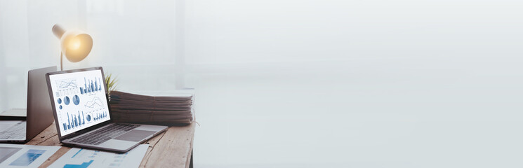 depicts a workspace with an open laptop showing graphs, a lit lamp, and documents on a wooden desk.