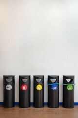 Frontal View of Five Color-Coded Recycling Bins Aligned on a Blue Floor Against a White Wall in...