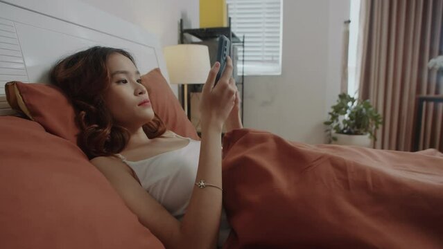 Handheld shot of young Asian woman using smartphone while resting in bed at weekend