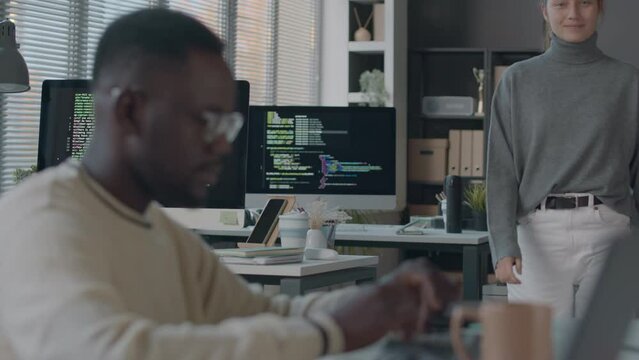 Medium shot of young Caucasian female programmer finishing coding on pc, getting up from her workplace and starting talking with Black male colleague, working together in modern office