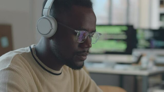 Tilt up shot of young Black male software developer typing on laptop keyboard while sitting at desk in modern office wearing wireless headphones and eyeglasses