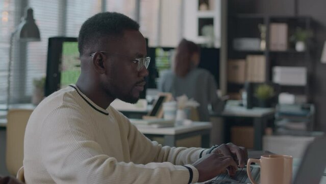 Waist up of young Black male software developer typing on wireless keyboard while co-working in modern programmers office with colleagues