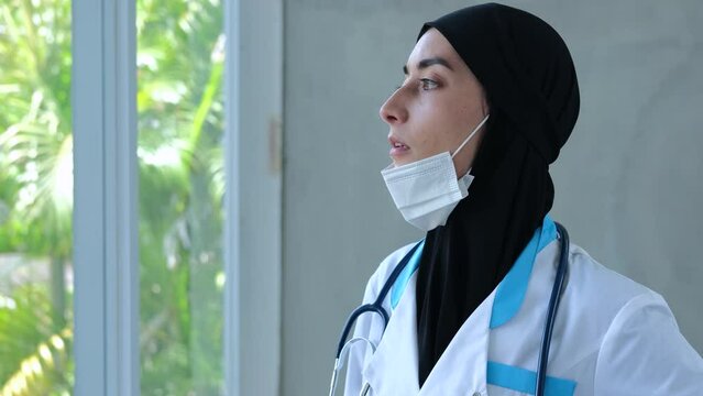 Muslim woman doctor at window takes off medical mask and rubs temple. Muslim woman doctor in black hijab sighs, pulls off medical mask and wearily rubs temple. Concept: tired doctor needs rest