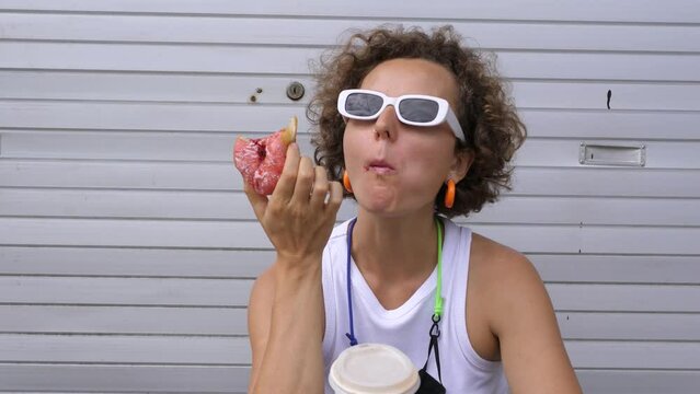 Lady eats snack donut with coffee. Close-up woman in white T-shirt and white sunglasses happily eats donut snack, licks lips and washes it down with coffee. Concept of snacking outdoors under sun