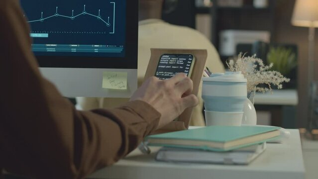 Back view of software engineer looking at chip interface on computer screen and program code on smartphone while working at desk in modern office