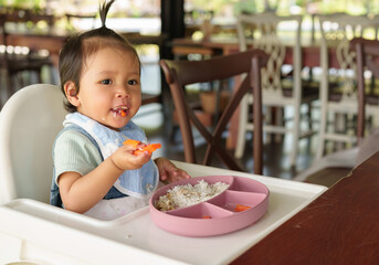 toddler girl eating food and vegetable by self feeding BLW or baby led weaning on highchair in...