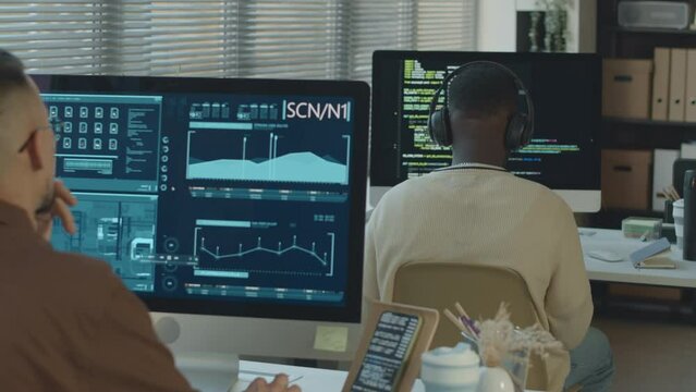 Back view of two ethnically diverse male software engineers examining chip interface and coding on computers while working together in modern programmers office