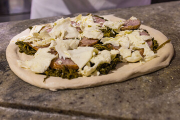 Pizza tradizionale napoletana condita con friarielli, salsiccia e mozzarella poggiata sul banco da lavoro in marmo e pronta per essere infornata 