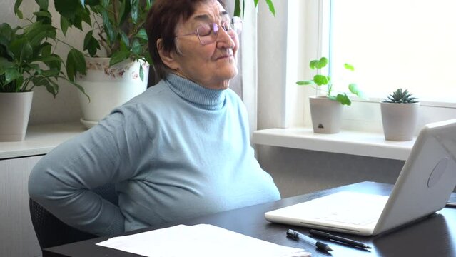 An elderly woman working at home on a computer experiences back pain. She takes a break, stretches, and massages her back to ease the discomfort. Maintaining health during long-term computer use.