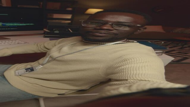 Vertical handheld POV of young African American male network engineer recording video of himself program coding in dark office at night