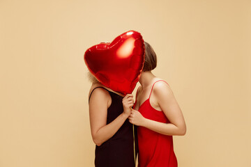 Two girls, lesbian couple in dresses kissing behind air balloon in heart shape over light beige background. Concept of lgbt community, love, Valentine's day, freedom, acceptance, February 14th - obrazy, fototapety, plakaty