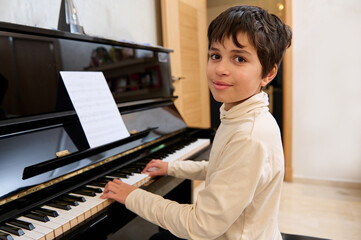 Confident handsome teen boy sitting at piano, smiles looking at camera, playing grand piano during...