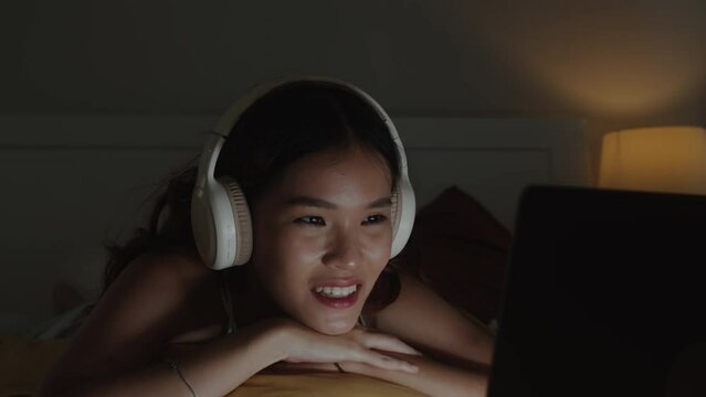 Medium close up of cheerful Asian girl watching movie on wireless computer using headphones lying on bed at home at nighttime