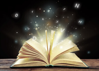 Open book with magic light and glowing letters flying out of it on wooden table against black...
