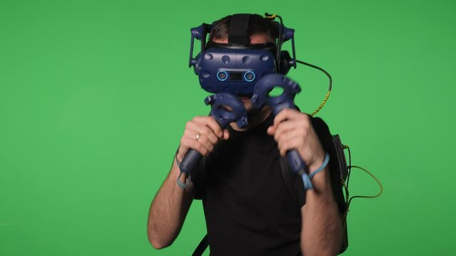 Portrait of a young man in virtual reality glasses with a joystick on a green screen background. A male gamer is playing virtual boxing. Chroma key.