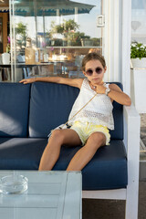 Ritratto di una bambina di nove anni seduta su di una poltrona con occhiali da sole e capelli legati.