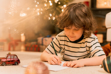 dark hair boy writes a letter with wish list to Santa Claus using color pencils laying on floor near Christmas tree.