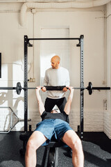 Unrecognizable sportsman on bench exercising with barbell with adult man instructor