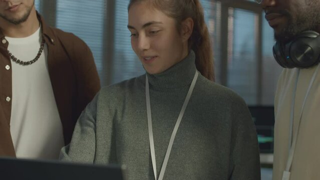 Tilting up shot of young Caucasian female IT coordinator pointing at computer screen while speaking to diverse male network engineers, working together in modern office in evening