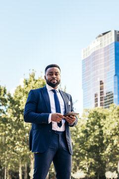 Low angle portrait of a black business man in a suit using a tablet outside and looking at camera smiling