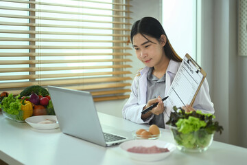 Female nutritionist showing clipboard in front of laptop explaining details of slimming program to...