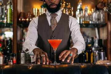 Cutout of a black bartender offering a cocktail served in a martini glass in a traditional cocktail bar - obrazy, fototapety, plakaty
