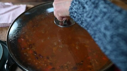 Woman's hand takes a lid off a large pot and stirs a freshly cooked bean soup that ist simmering