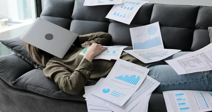 Tired woman sleeps on the couch with documents and laptop. Scattered document with text and graphics and tired freelancer