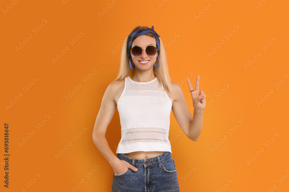 Wall mural Smiling hippie woman showing peace sign on orange background