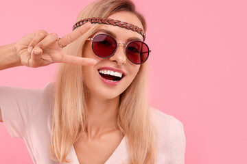 Portrait of smiling hippie woman showing peace sign on pink background. Space for text