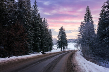 asphalt road through forested countryside in winter at sunset. trees and hills in snow. dramatic...