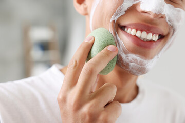 Happy young man washing off face mask with sponge in bathroom, closeup
