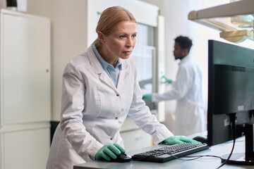 Medium shot of mature female laboratory technician wearing white coat and gloves standing at desk...