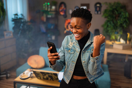 Happy african american teen girl lady woman with short hair using cell phone holding in hands smiling celebrating raising fists.